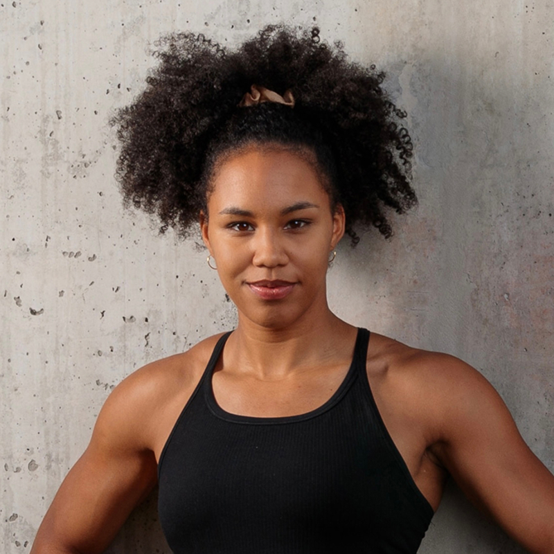 Confident young woman with curly hair wearing a black tank top standing against a textured concrete wall.